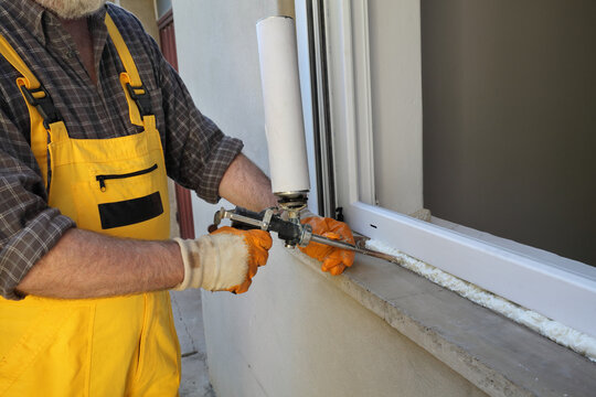 Worker installing new plastic window, using handgun with polyurethane foam 