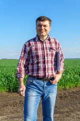Fototapeta premium a man as a farmer poses in a field, dressed in a plaid shirt and jeans, checks reports and inspects young sprouts crops of wheat, barley or rye, or other cereals, a concept of agriculture and agronomy