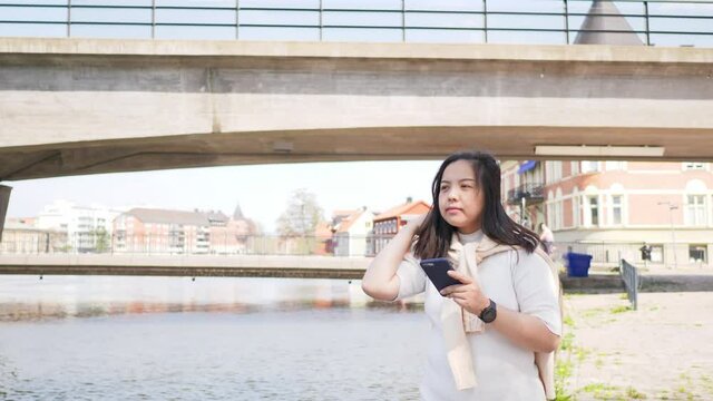 Asian black straight hair woman standing by a river in town and using her smartphone to text to her friend, having a great day on free time. Enjoy life and take a deep breath