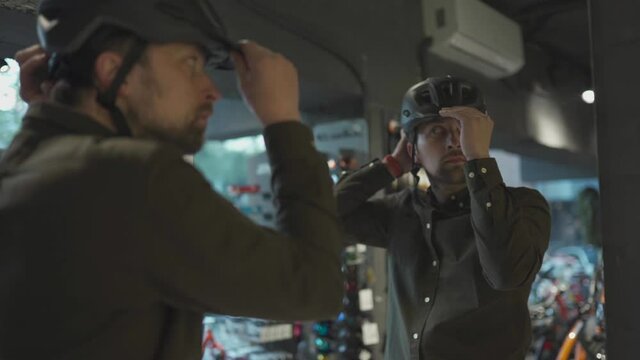 Young man came to the bicycle store. He is measuring the helmet. Male chooses helmet in sports equipment store. Purchase of new sports helmet. Customer with bicycle helmet trying on near the mirror