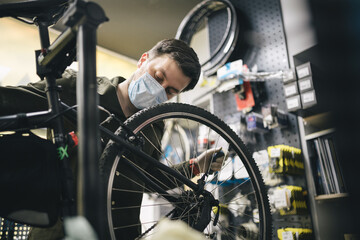 Bicycle mechanic wearing protective mask and gloves repairs customers bicycle wheel in accordance with quarantine standards during coronavirus pandemic. Small business during covid 19 lockdown