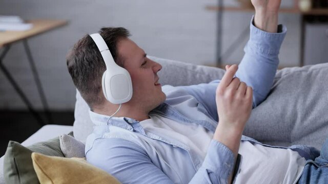 Caucasian male enjoying music on the couch at home. Young man listening to music in modern white headphones. Happy guy dancing and listening to music with headphones. People lifestyle