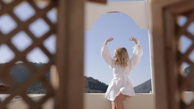 Blonde girl enjoying summer and hiding from the sun in white bungalow. Mountains and sea on background. Vacation holidays at luxurious beachfront hotel resort.