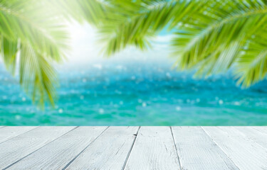Empty white wooden table on blurry beautiful nature green palms leaf on tropical beach with bokeh sun light wave abstract background. Copy space of summer vacation and business travel concept.