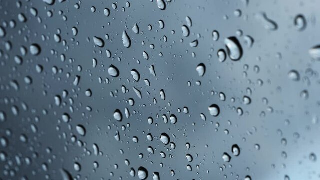 Water raindrop on a clear window glass. Raining on a car windscreen on a moody day. Moving droplets close up in front of a dark cloudy sky. Liquid fluid splashing on the windshield.
