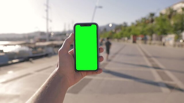 POV Close-up young man watching on iPhone 11 green mock-up blank screen browsing social network in smartphone walking outdoors in the marina bay.