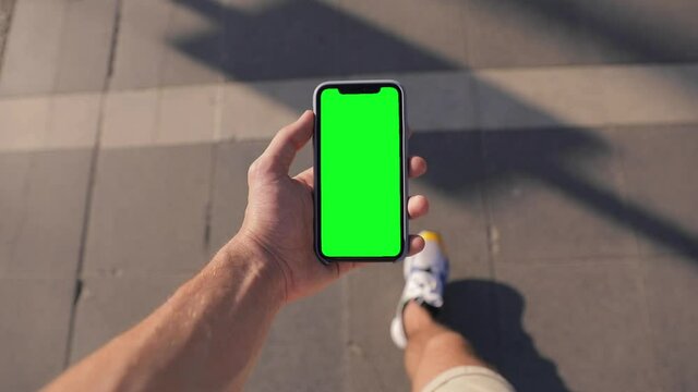 Closeup of a man's hand holding an Apple iPhone 11 Pro mobile phone with a vertical green screen. Walking on the summer street. Promenade