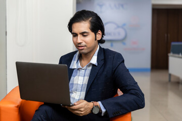 Portrait of young successful Indian business man in dark suit sitting on couch and working on laptop, Corporate environment.