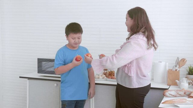 Young asian mother and son fat standing eating apple in the kitchen at home, fruits for healthy, mom and boy dieting and weight loss, female and child with overweight and nutrition, plus size.