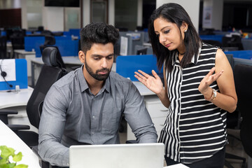 Portrait of two young serious businesspeople looking at laptop and discussing project at workstation, corporate environment.