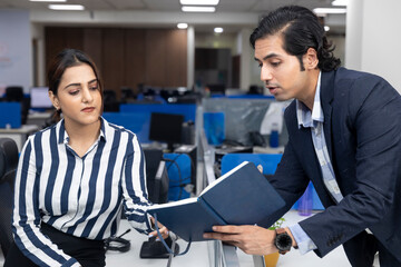 Portrait of two young Indian businesspeople sharing diary and discussing project at workstation, corporate environment.