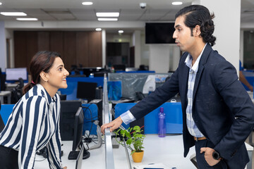 Portrait of two young Indian businesspeople discussing project across workstation, corporate environment.