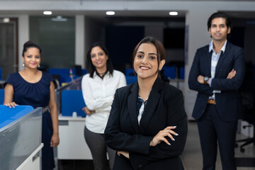 Confident Indian businesswoman standing infront of her office colleagues, selective focus, corporate environment, team members, business office.