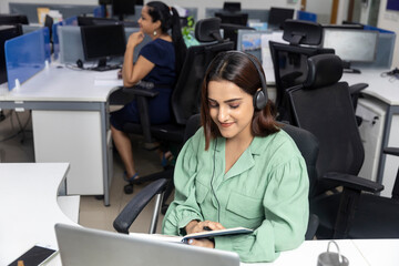 Portrait of pretty Indian businesswoman wearing headphones, sitting at her workstation against...