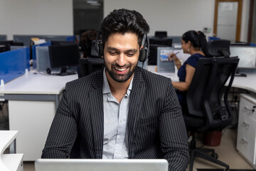 Portrait of young Indian businessman wearing headphones, sitting at his workstation against office...