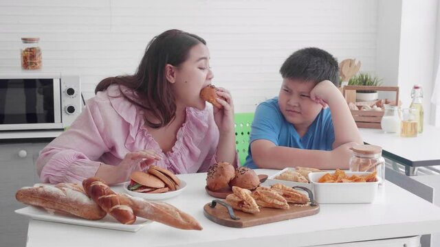 Asian family relationship of son upset with mother fat eating bread in the kitchen at home, mom hungry and eating bun with unhealthy food, two people, woman overweight, health care concept.