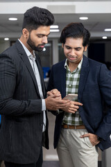 Portrait of two young handsome Indian businessmen looking into phone, discussing project, corporate environment.