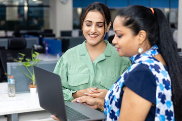 Portrait of two young Indian businesswomen looking into laptop, discussing project, corporate environment, office background.