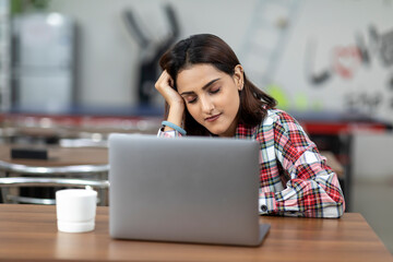 Portrait of a tired Indian woman working on her laptop, sitting in an office cafeteria, coffee shop, casual work environment, rubbing her forehead.