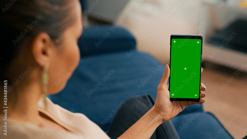 Poster caucasian female sitting on sofa in living room while socializes on internet app using phone with mo