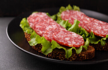 Delicious sandwich on black grain bread with lettuce and smoked sausage on plate on black background