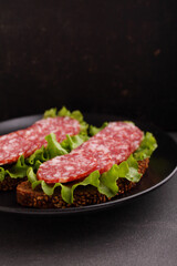 Delicious sandwich on black grain bread with lettuce and smoked sausage on plate on black background