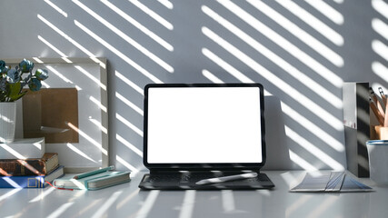 Computer tablet, notebook and pencil holder on white desk near window with sunlight.