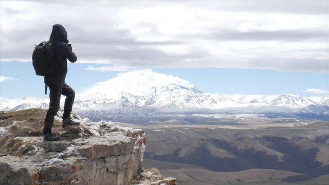 a male tourist traveler on a hill overlooking the mountain range of the Caucasus mountains Elbrus stands boldly on a rock and takes pictures on his phone