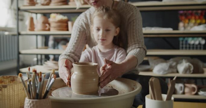 Grandmother teaches her granddaughter working on a pottery rotating wheel. Senior woman and cute little girl make pot on a spinning pottery wheel. Hobby, leisure, arts and crafts concept.