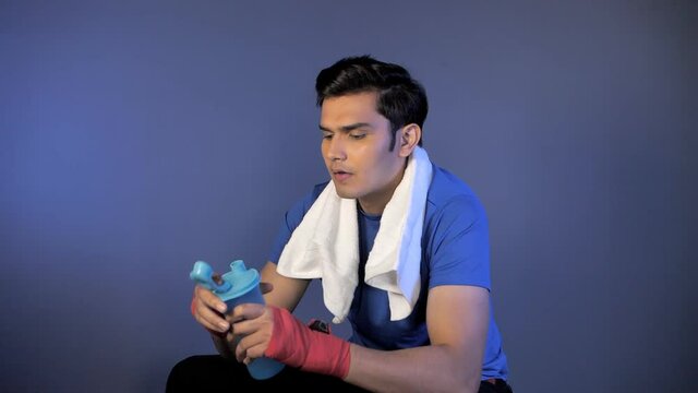An athlete with gloves and towel drinking health drink from sipper - sitting against a dark background. Young Indian man drinking water while relaxing after his daily workout session - healthy acti...