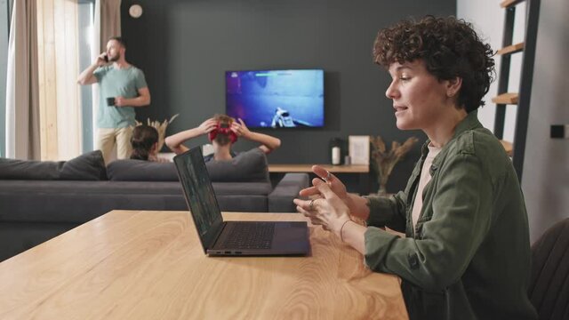 PAN Medium Shot Of Confident Woman Having Business Video Call On Laptop From Home While Her Husband Talking On Phone Standing By Window And Two Kids Playing Video Games In Background