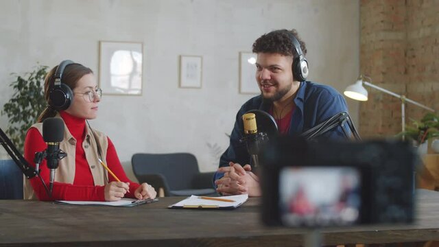 Cheerful young man in headphones sitting in studio and giving interview to female broadcaster as digital camera recording video