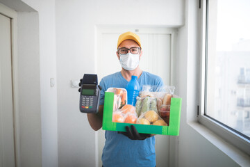 Fototapeta na wymiar Delivery guy in front of home door with protective mask holding box with groceries and POS for contactless payment.