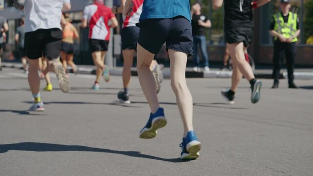 Slow Motion Feet Of Athletes Running Marathon In City, Police Officer And Onlookers On Roadside. Partial View From Behind Of Contestants Of Sports Event. Healthy Lifestyle. Concept Of Sport