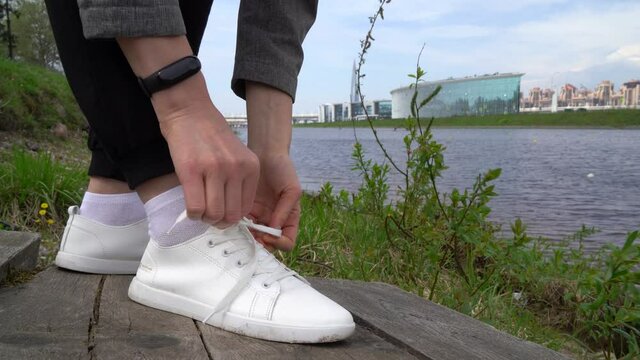 A man ties his shoelaces on white sneakers on the street against the background of the river and nature close-up.