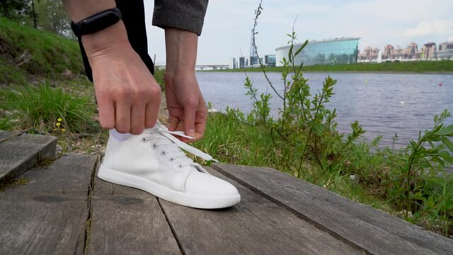 A man ties his shoelaces on white sneakers on the street against the background of the river and nature close-up.