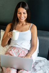 Beautiful woman sitting on bed, holding in hands cup of coffee and using laptop computer