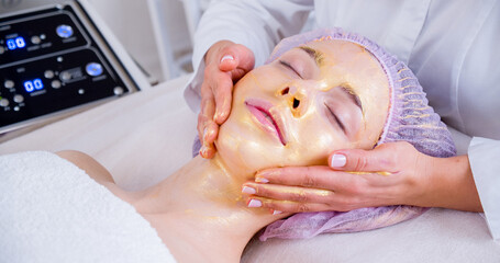 Obraz na płótnie Canvas Cosmetologist applies a woman a therapeutic mask on her face. Woman in a spa salon on cosmetic procedures for facial care. Beautician makes a massage of the face with a moisturizing mask on a face.