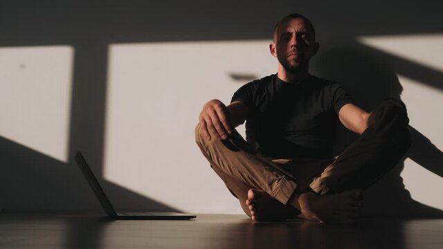 A man is sitting in a lotus position in a room With a laptop on the floor A shadow falls from the window A break in work Concentration Slow motion