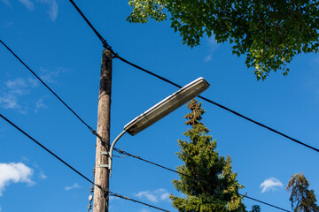 A street lamppost with wires next to green trees on a street. Wooden pole with a lantern and...
