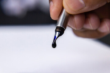 Drop of blue ink dripping out from a fountain pen nib over white paper.