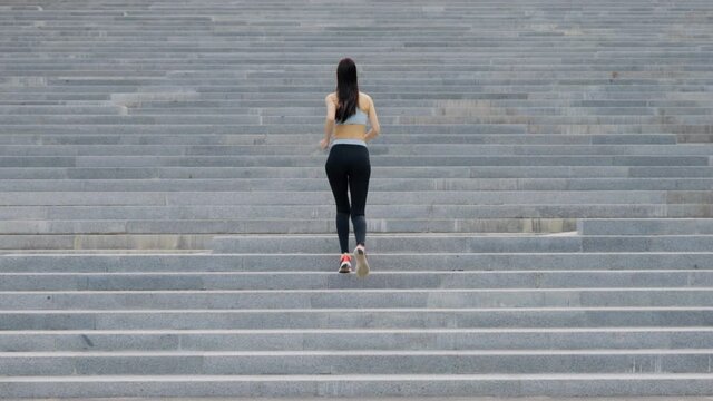 Woman with ponytail wearing sportswear running up on staircase outside. Back view athletic female jogging in urban setting. Healthy lifestyle. Concept of fitness