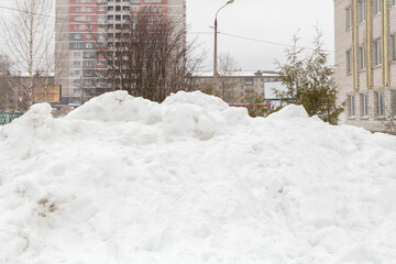 A large snowdrift on a city street with high-rise buildings. A block of snow with dirty lumps lie in the city in the courtyard of an apartment building. Gloomy winter day.
