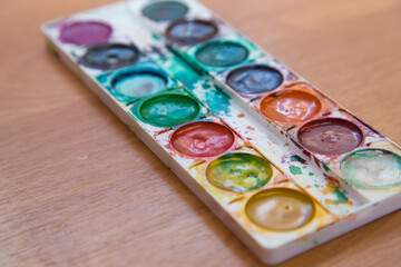 On the table is an old set of multicolored watercolor paints for painting. Top view of half-empty jars of watercolors. Flat layout on the table.