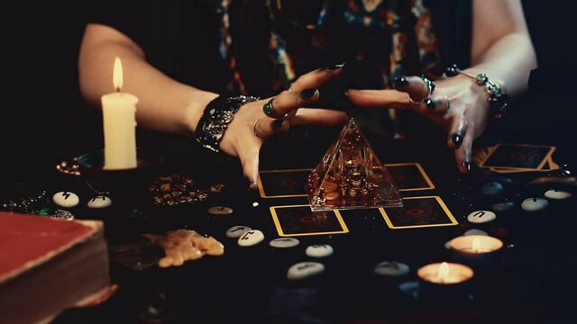 Divination and magic. The fortune teller telling fortunes on the glass pyramid with the cards. Close-up of hands with dark manicure and rings. Black table with burning candles.
