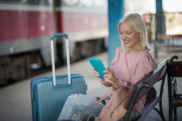 Smiling blond woman  on train station using digital tablet. - obrazy, fototapety, plakaty