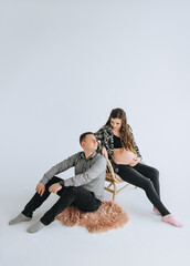 Stylish man and a beautiful pregnant woman with a big belly sit on a wicker wooden chair and on a woolen rug on a white background cyclorama, photo studio and tenderly hug. Photography, concept.