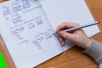 A girl's hand with a ballpoint pen close-up writes numbers in a notebook. The student completes the task. A student solves examples in mathematics at school at a desk. Warm soft daylight.