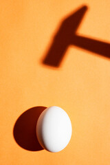 Minimal concept with an egg under the harsh light and a hammer shadow on an orange background