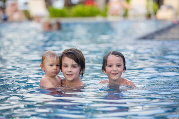 Adorable happy little child, toddler boy, having fun relaxing and playing with his older brothers in a pool on sunny day during summer vacation